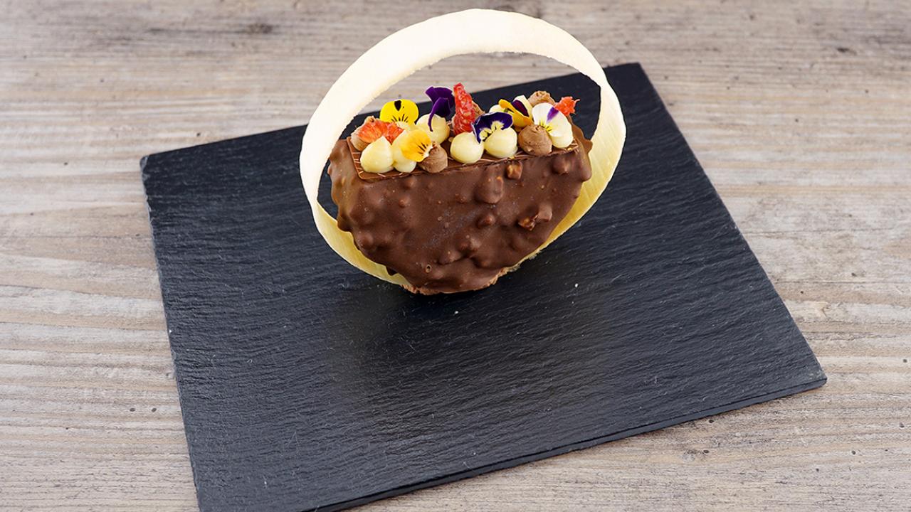 A chocolate-covered dessert with nuts, topped with colorful edible flowers, sits on a black slate plate, with a curved white chocolate decoration around it.