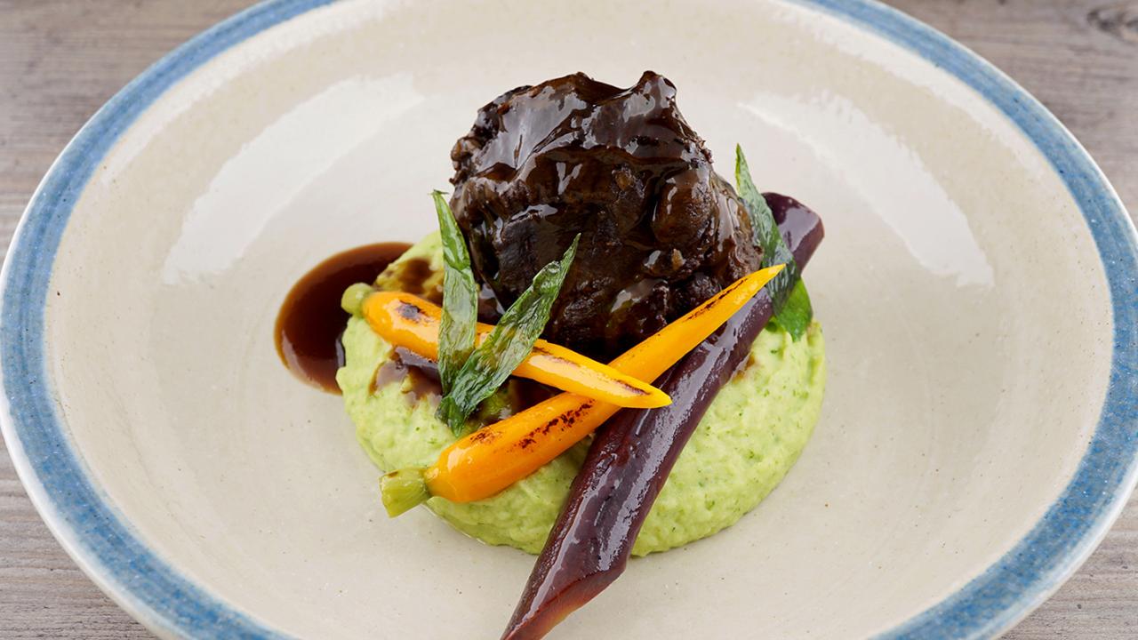 A dish with braised meat on pea purée, garnished with baby carrots and asparagus spears, drizzled with dark sauce.