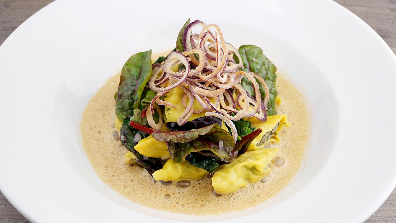 A gourmet dish featuring pasta, leafy greens, and crispy fried onions, served on a bed of creamy sauce on a white plate.