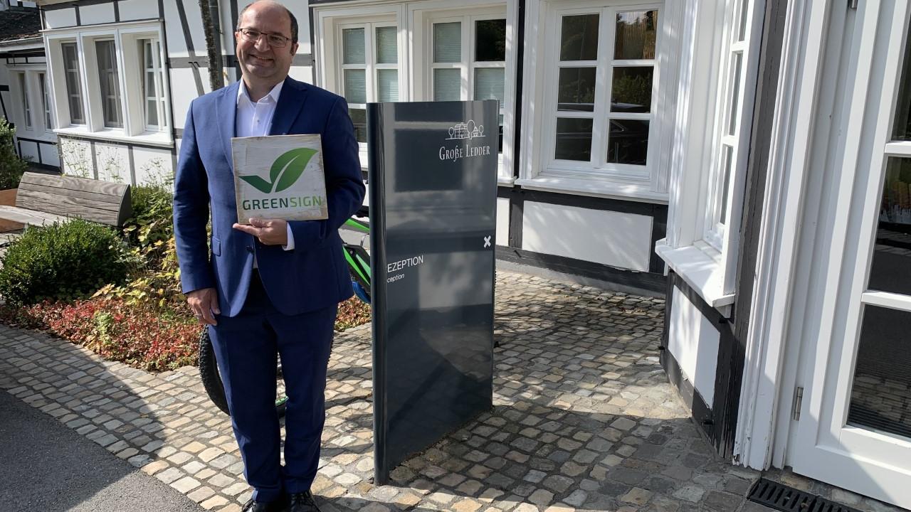Man in a blue suit holding a document with a green leaf design, standing outside a building with a sign labeled "Reception".