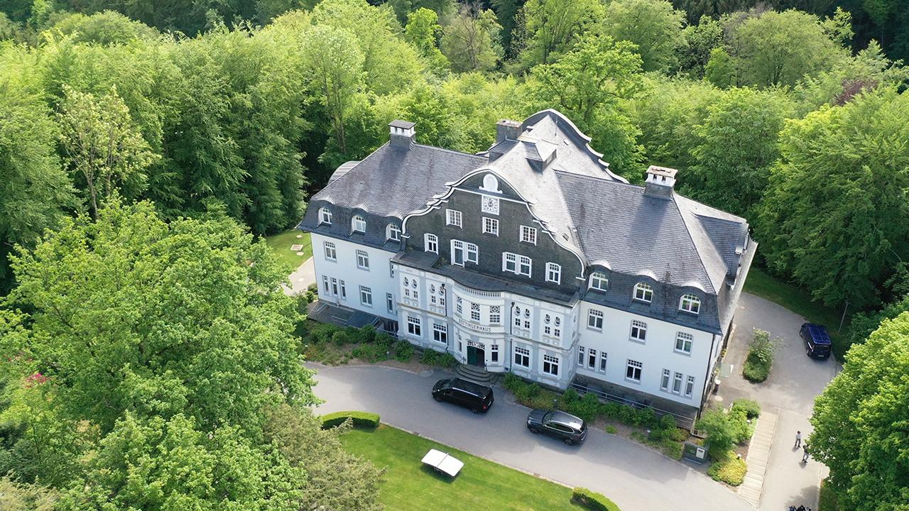 Aerial view of a large, grey-roofed building surrounded by dense green trees. Several cars are parked on the driveway, and a few people are visible near the entrance.