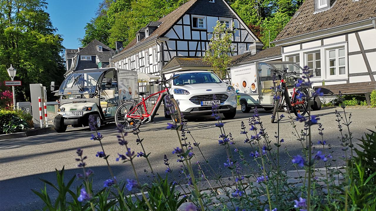 A street scene with a white car, two small delivery vehicles, and two bicycles. Traditional half-timbered houses and trees are in the background, with purple flowers in the foreground.
