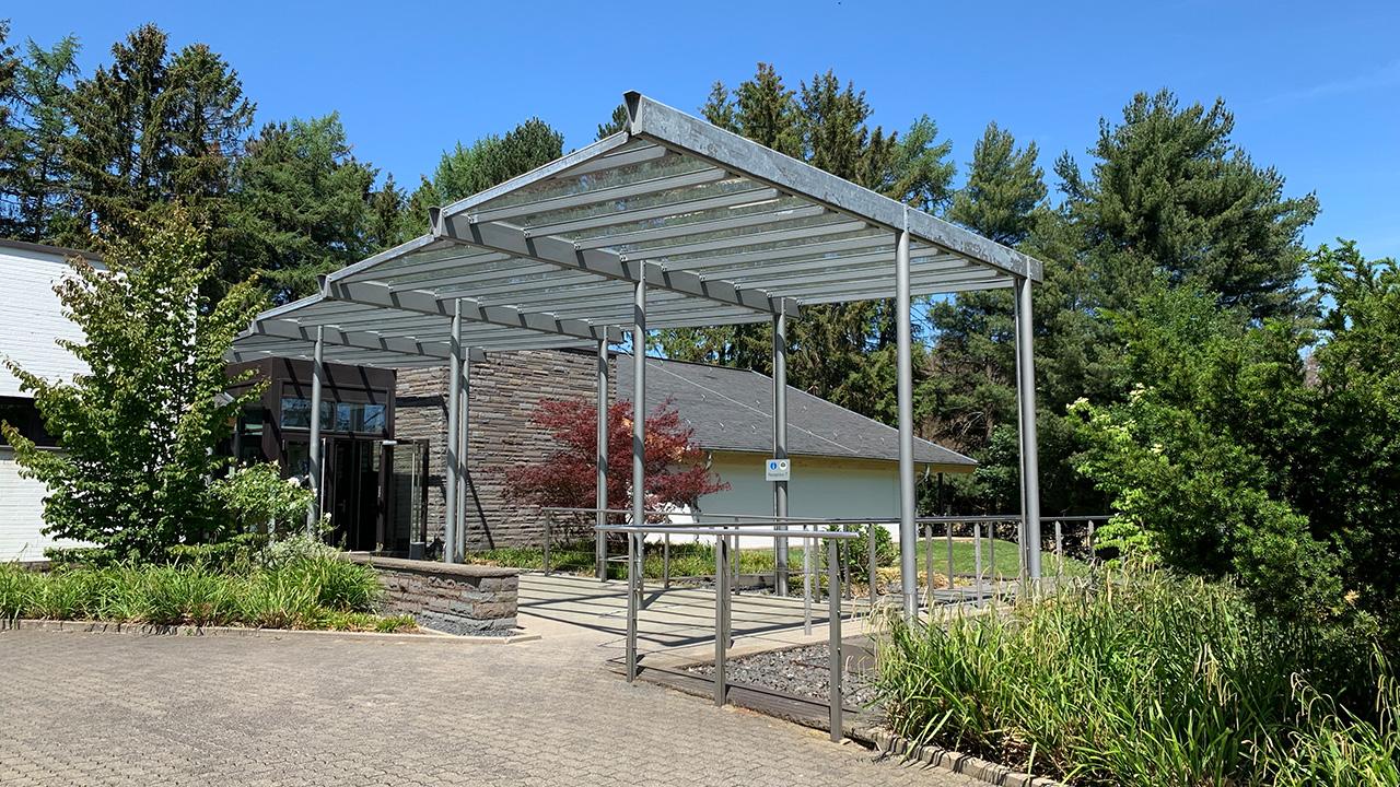 A metal pergola stands on a brick pathway surrounded by greenery and trees, with a building in the background.