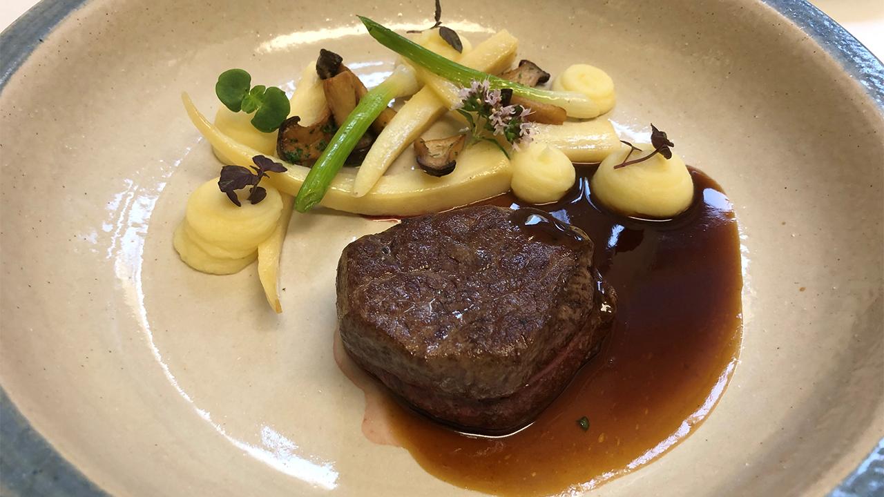 A gourmet dish featuring a piece of seared meat with sauce, accompanied by purée, sautéed vegetables, and garnished with herbs on a ceramic plate.