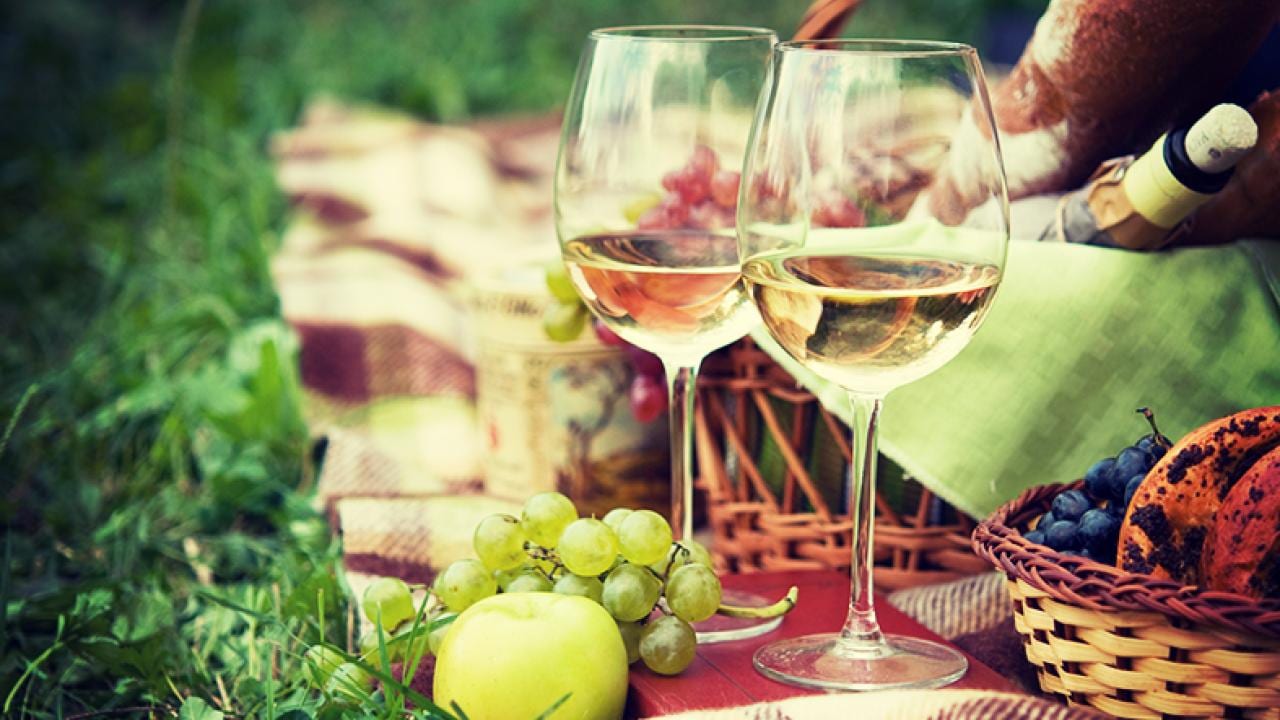 Two wine glasses, grapes, an apple, bread, and a wicker basket on a picnic blanket outdoors.