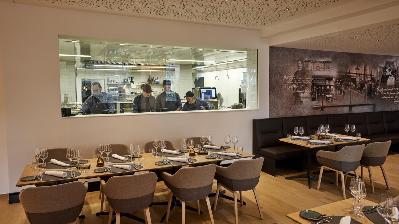 A modern restaurant interior with neatly set tables in the foreground and chefs working in an open kitchen behind a glass window.