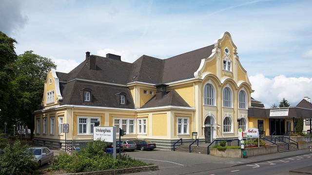 Yellow building with decorative facade, large arched windows, and a dark roof. Several cars are parked outside. Signs are visible in the foreground near a road.