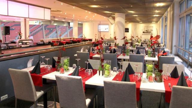 A modern restaurant interior with tables set for dining, featuring red and black accents, neatly folded napkins, and decorative centerpieces in glass vases. Large windows line the right side.