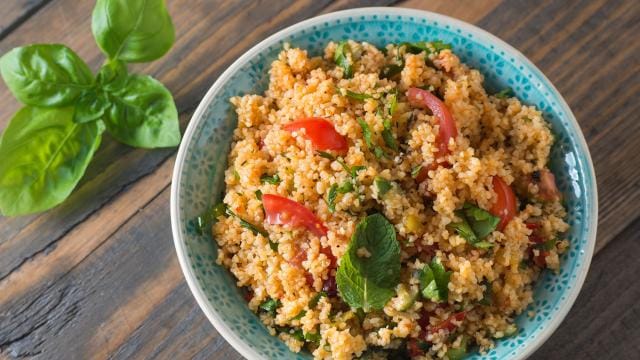 A bowl of couscous salad with tomatoes, herbs, and mint leaves, on a wooden table. Fresh basil leaves are placed nearby.