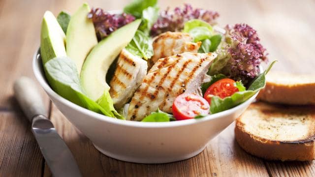 A white bowl with grilled chicken, avocado slices, cherry tomatoes, and mixed greens on a wooden table next to toasted bread and a knife.