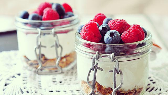Two glass jars filled with layered granola, yogurt, and topped with fresh raspberries and blueberries on a lace-patterned surface.