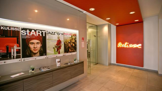 Modern lobby with a red ceiling and a neon "Kulisse" sign on the wall, featuring promotional posters and a reception counter with brochures.