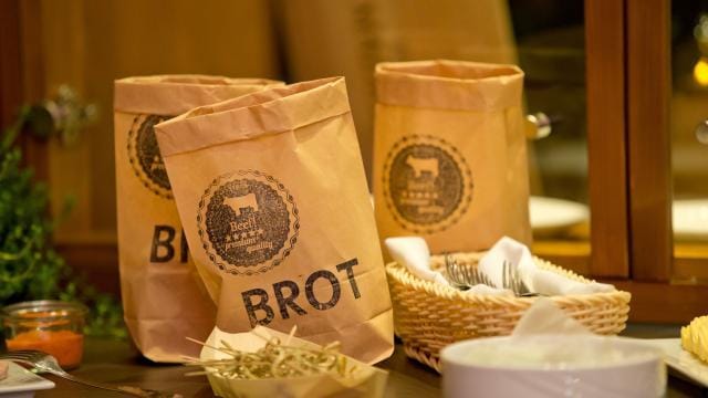 Brown paper bags labeled "BROT" on a table with plants, a basket of utensils, and small dishes in the foreground.