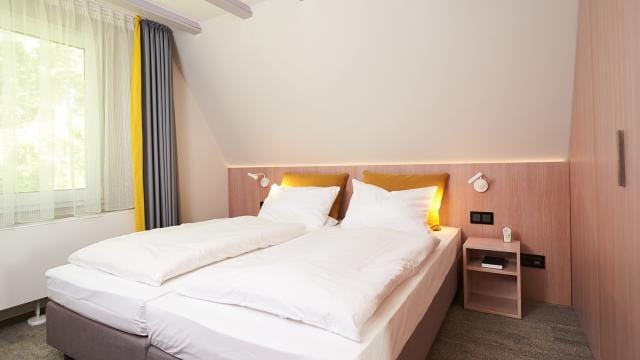 A modern bedroom with two single beds pushed together, white bedding, wooden nightstand, wall-mounted lamps, and a window with sheer and gray curtains.