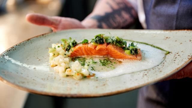 A person holds a plate with cooked salmon, topped with herbs and a white foam sauce, next to a small portion of diced vegetables.