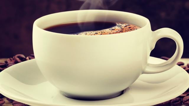 A steaming cup of black coffee sits on a white saucer, surrounded by scattered coffee beans.