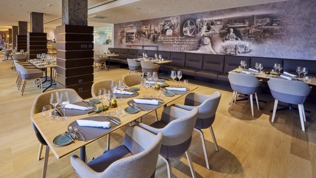 Modern restaurant interior with wooden tables, beige chairs, set with glasses and napkins. Background features a large mural.
