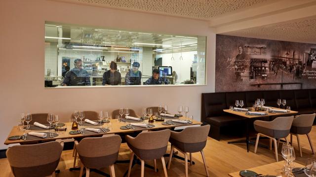 Empty restaurant dining area with set tables and chairs. Chefs are visible working in the kitchen through a large glass window.