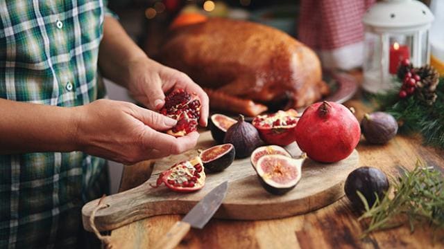 A person prepares pomegranates on a wooden board, with figs and a roast turkey in the background.