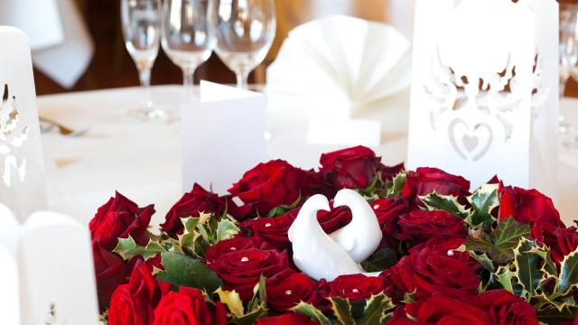 Tischset mit Gläsern, weißen Besteckhaltern und einem Tafelaufsatz aus roten Rosen mit zwei Porzellanschwänen, die ein Herz bilden.