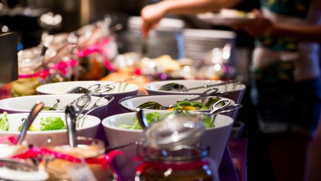 Ein Buffet mit verschiedenen Salaten und Toppings in weißen Schüsseln. Im Hintergrund serviert eine Person Essen.