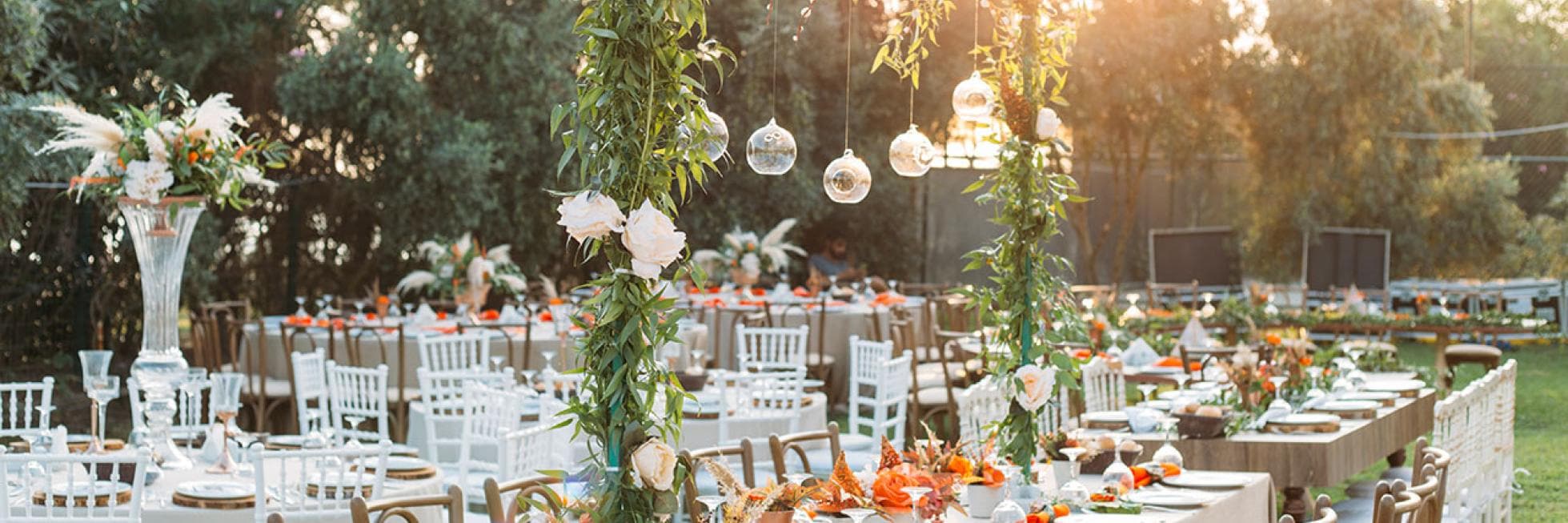 Outdoor wedding reception setup with long tables, white tablecloths, and floral centerpieces. Greenery and hanging glass ornaments adorn the area. Sunlight filters through surrounding trees.