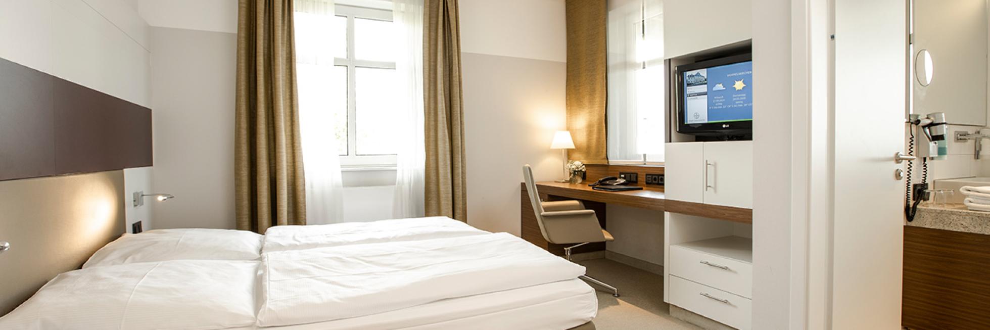 A modern hotel room with a double bed, desk, wall-mounted TV, and a glimpse of a bathroom. Beige and white decor with natural light from a window.