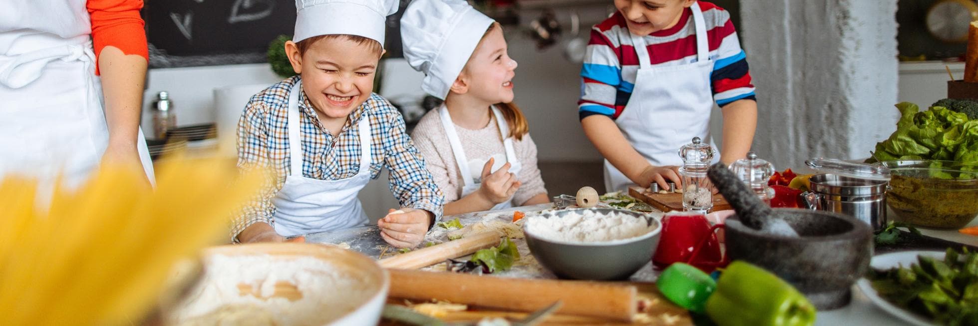 Three children wearing chef hats and aprons are smiling and preparing food in a kitchen. Various ingredients and utensils are on the table.