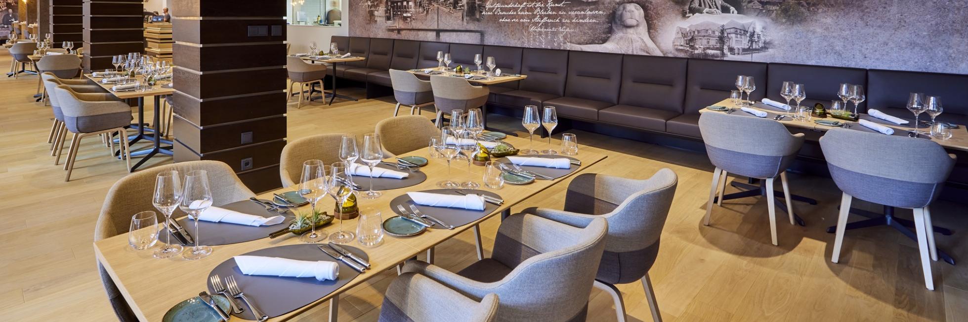 Modern restaurant interior with wooden tables, beige chairs, set with glasses and napkins. Background features a large mural.