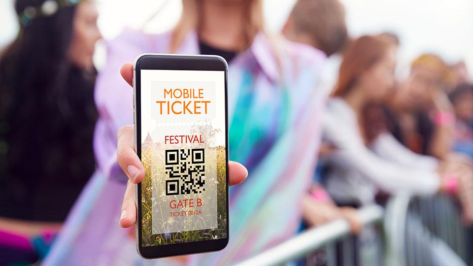 A person holds a smartphone displaying a mobile ticket with a QR code for a festival at Gate B. People and a fence are visible in the background.